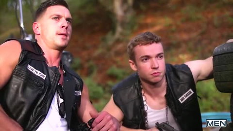 Two muscular men in leather vests holding guns in outdoor setting