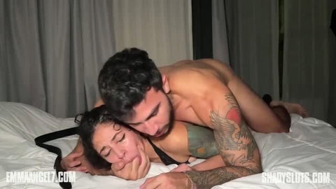 Man dominating woman in bed, hand on face, tattooed couple sex scene