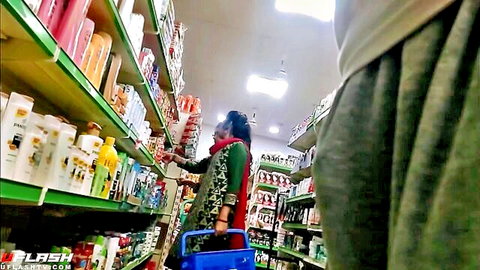 Indian woman in green salwar kameez reaches for shelves, eyeing bulge in store