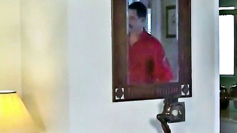 Man in red shirt reflected in wooden mirror in cozy apartment room with lamp.