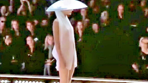 Slim runway model struts in sheer white dress and oversized hat amid cheering crowd