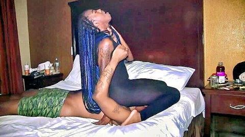 Tattooed ebony babe with blue braids straddles black stud's crotch in shiny black bodysuit on bed