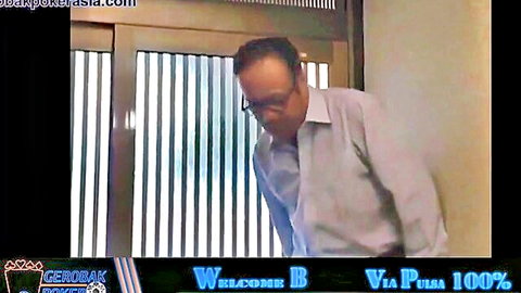 Clothed Asian man in glasses and white shirt peers through vertical blinds in office