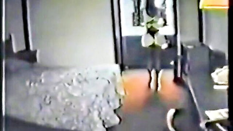Amateur wife in sheer white bridal gown standing by bed in hotel room teasing