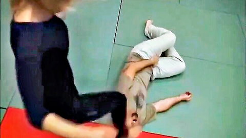Blonde woman dominates man in wrestling submission hold on gym mat