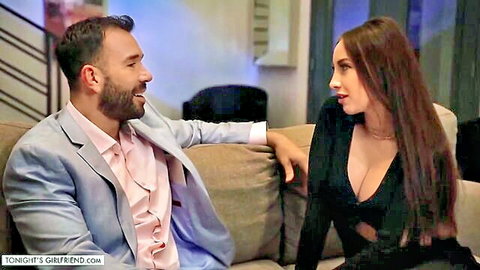 Busty brunette in deep cleavage black dress flirts with suited bearded man on couch
